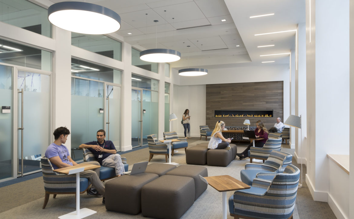 Multiple employees in a flexible space, with chairs, ottomans, tables, lamps, lights and a fireplace