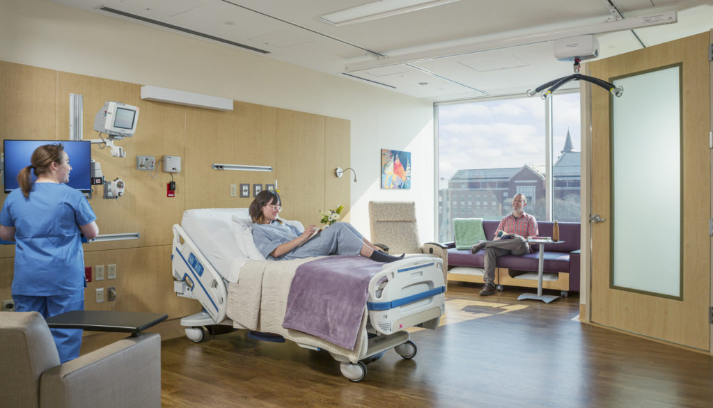 Hospital room with patient on bed and nurse talking to visitor sitting on couch