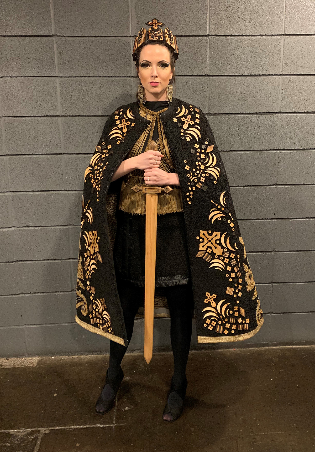 Model wearing black and tan design competition cape, shirt and crown holding wooden sword