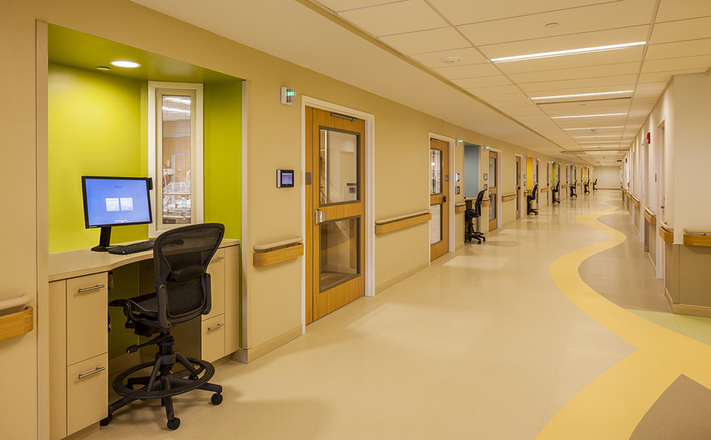 Long hospital hallway with workstations and doors along wall with colorful floors and alcoves 