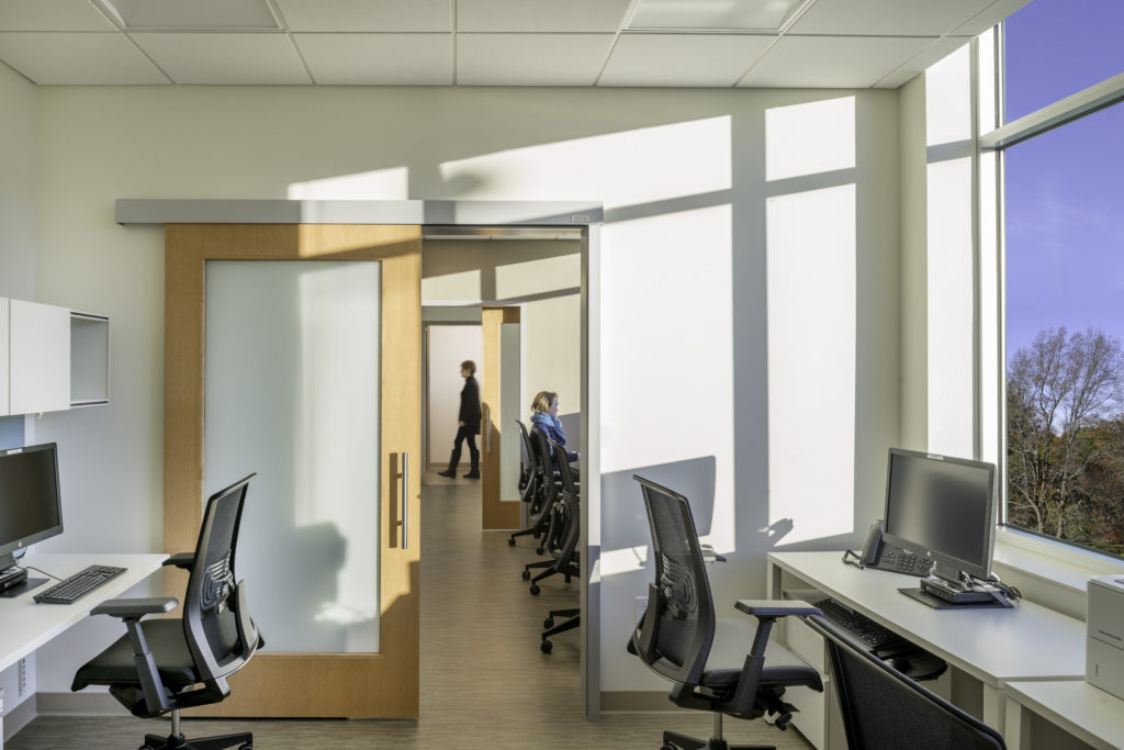 Brightly lit office workstations with one woman working and one woman walking in hallway