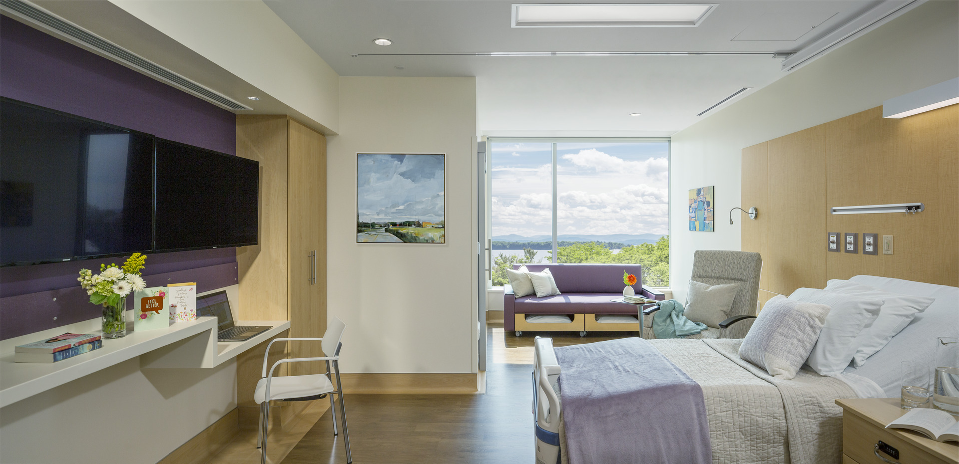 Patient room wth television, desk, laptop, chairs, couches, books, flowers and a bed, with paintings on the walls