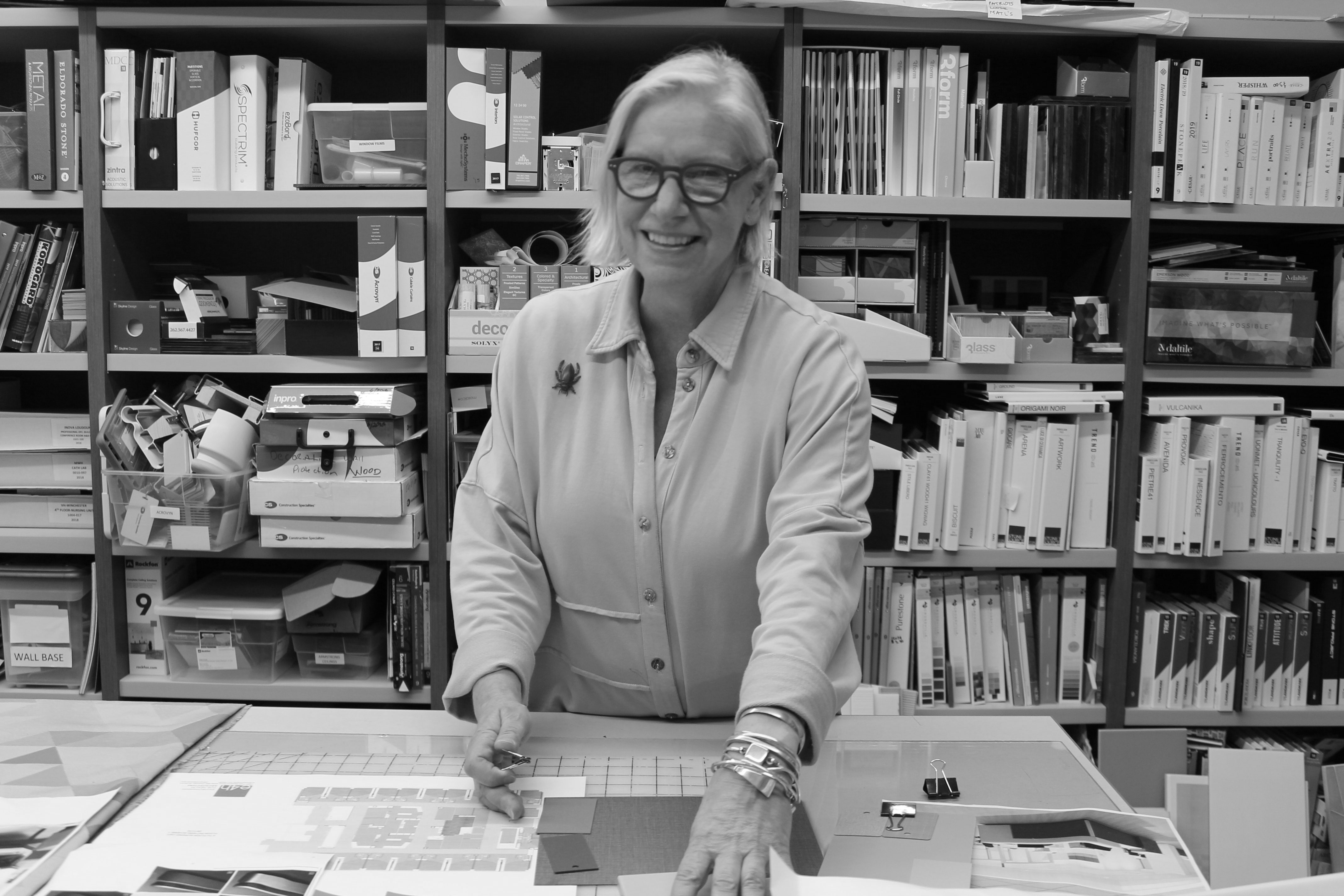 Nan Schramm, Senior Associate, wears a button-down top, metal bracelets, glasses, and smiles while working on a design at her workstation