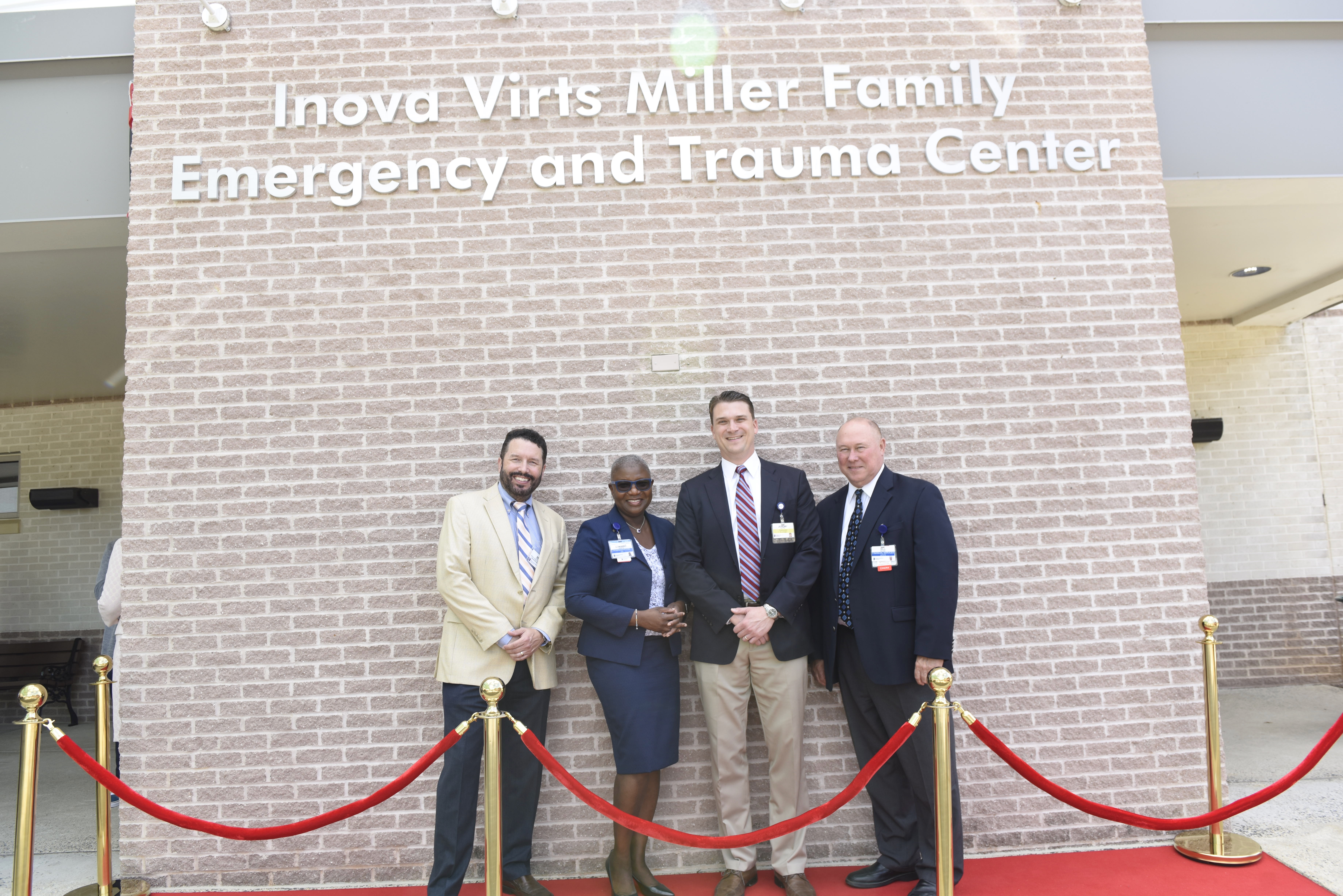 Inova Virts Miller Family Emergency Trauma Center ribbon cutting - Four people dressed in business attire smile while standing on a red carpet in front of a brick wall